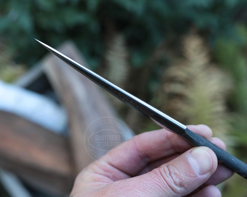 May include: A close-up of a metal awl with a sharp, pointed tip. The awl has a dark handle and a silver-colored blade. The tool is held in a hand against a blurred green background. The text "SWAYTOBOR TOOLS" is visible.