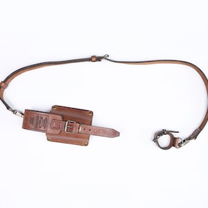 Puede incluir: Una funda de teléfono de cuero marrón con una correa y una hebilla. La funda tiene un pequeño bolsillo en la parte delantera con el texto "The Original" impreso en él.