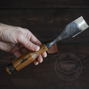 以下が含まれることがあります： 木製のハンドルと金属製の刃を持つ木彫りのノミ。刃はわずかに湾曲しており、ハンマーで仕上げられています。ハンドルは木でできており、金属製のバンドが巻かれています。ノミは手で持たれています。