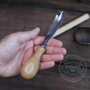 May include: A close-up of a wood carving tool with a wooden handle and a metal blade. The tool is held in a person's hand. The tool is labeled "Strongway Tools" and "Handmade Forge of Britain".