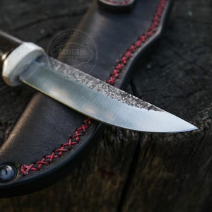 May include: A handcrafted knife with a dark wooden handle and a silver blade, resting in a black leather sheath. The sheath features red stitching and a decorative eyelet. The knife is designed for outdoor use.