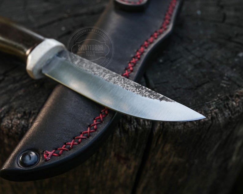 May include: A handcrafted knife with a dark wooden handle and a silver blade, resting in a black leather sheath. The sheath features red stitching and a decorative eyelet. The knife is designed for outdoor use.