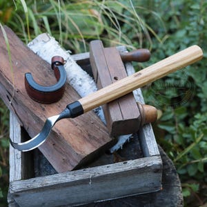 Könnte beinhalten: Ein Holzbearbeitungswerkzeug-Set mit einem gebogenen Ziehmesser mit Holzgriff, einem Lederriemen und einem Hobel aus Holz. Die Werkzeuge werden in einer rustikalen Holzkiste vor einem Hintergrund aus Grünzeug präsentiert.