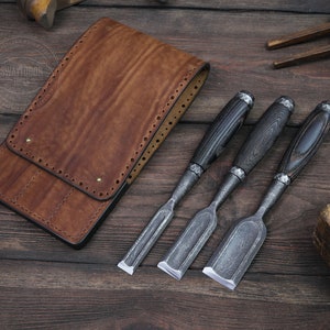 May include: A set of three wood carving chisels with black wood handles and a brown leather tool roll. The chisels are made of steel and have a distressed finish.