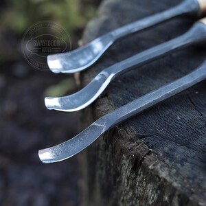 Może przedstawiać: Zbliżenie trzech metalowych narzędzi do rzeźbienia z drewnianymi uchwytami. Narzędzia mają zakrzywione ostrza i są ułożone na teksturowanej drewnianej powierzchni. Narzędzia są prawdopodobnie używane do obr&oacute;bki drewna lub rzeźbienia.