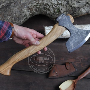 May include: A hand holds a handmade axe with a wooden handle and a dark grey metal head. The axe has a leather sheath and a wooden spoon is also visible in the image. The axe is labelled "Strongway Tools" with the text "Handmade Forge of Ukraine".