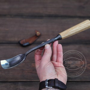 May include: A hand holds a black metal woodworking tool with a wooden handle. The tool has a curved blade and is used for carving wood. The tool is made by Strongway Tools, a company based in Ukraine.