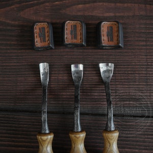 May include: Three wood carving tools with brown wooden handles and metal blades. The tools are arranged in a row on a dark brown wooden surface. The tools are labeled "Strongway Tools" in a white circle.