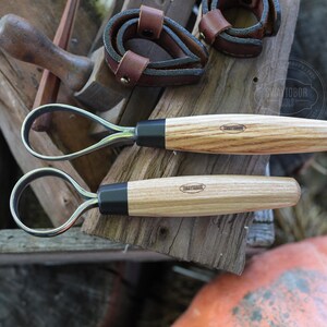 May include: Two hoof knives with wooden handles and black ferrules are displayed on a weathered wooden surface. The handles have a light wood grain pattern. The image also shows leather straps and other tools, suggesting a farrier or horse care theme.