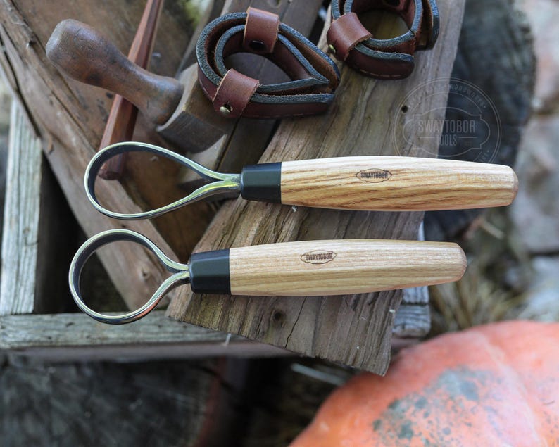 May include: Two hoof knives with wooden handles and black ferrules are displayed on a weathered wooden surface. The handles have a light wood grain pattern. The image also shows leather straps and other tools, suggesting a farrier or horse care theme.