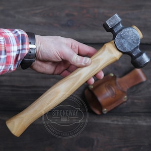 Könnte beinhalten: Ein Holzgriff-Kugelhammer mit einem schwarzen Metallkopf. Der Hammer wird in der Hand gehalten und hat eine Lederscheide.