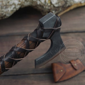 May include: A black and brown leather-wrapped axe head with a wooden handle. The axe is on a wooden surface with a leather sheath next to it. The axe has a Strongway Tools logo on the blade.