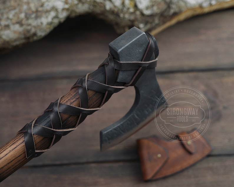 May include: A black and brown leather-wrapped axe head with a wooden handle. The axe is on a wooden surface with a leather sheath next to it. The axe has a Strongway Tools logo on the blade.