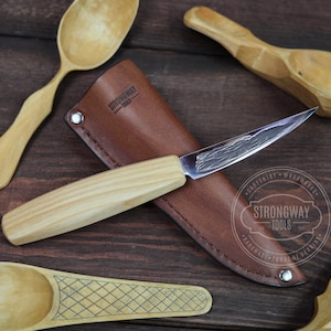 May include: A wooden handled carving knife with a leather sheath. The knife has a textured blade and the sheath has the text "Strongway Tools" embossed on it. The knife is lying on a wooden surface with wooden spoons.