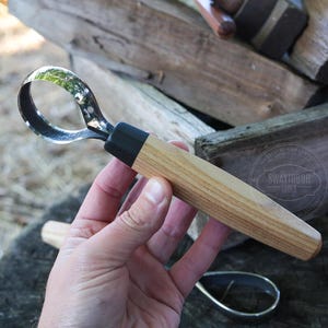 May include: A wood carving hook knife with a curved blade and a light brown wooden handle. The blade is made of metal and has a black section where it connects to the handle. The handle is cylindrical and has a black section near the blade.