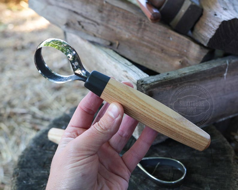 May include: A wood carving hook knife with a curved blade and a light brown wooden handle. The blade is made of metal and has a black section where it connects to the handle. The handle is cylindrical and has a black section near the blade.
