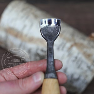 May include: A close-up of a wood carving tool with a sharp, three-pronged blade and a wooden handle. The tool is held in a hand, and the background is a piece of birch bark.