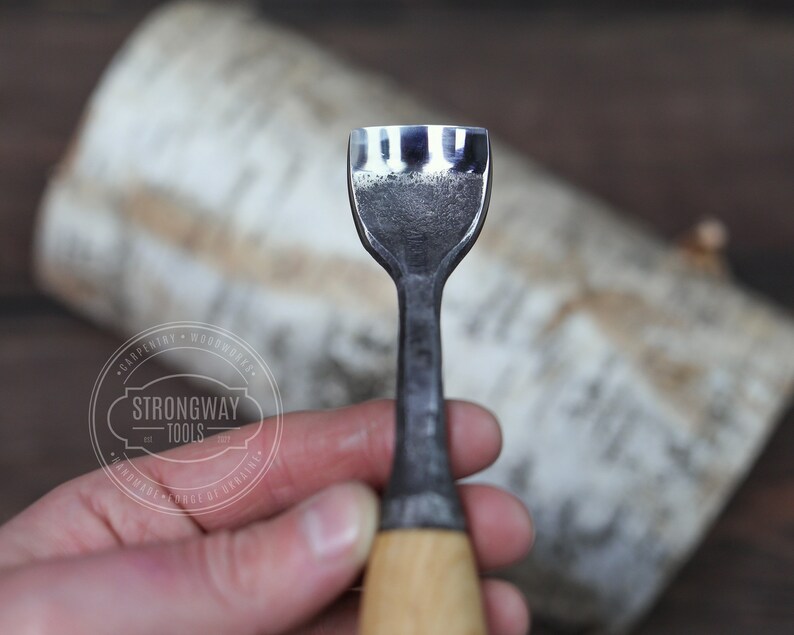 May include: A close-up of a wood carving tool with a sharp, three-pronged blade and a wooden handle. The tool is held in a hand, and the background is a piece of birch bark.