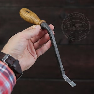 Op de afbeelding: Een hand houdt een klein, gebogen houtbewerkingsgereedschap vast met een houten handvat. Het gereedschap is gemaakt van metaal en heeft een scherpe rand. Het gereedschap is gelabeld met "Strongway Tools" met de tekst "Craftsman - Woodworks - Handmade - Forge of Berlin".