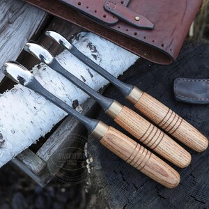 Może przedstawiać: Cztery dłuta do obr&oacute;bki drewna z polerowanymi metalowymi ostrzami i drewnianymi uchwytami. Uchwyty są jasnobrązowe z trzema ciemnobrązowymi paskami. Dłuta ułożone są na kawałku kory brzozy, z sk&oacute;rzanym etui na narzędzia w tle.