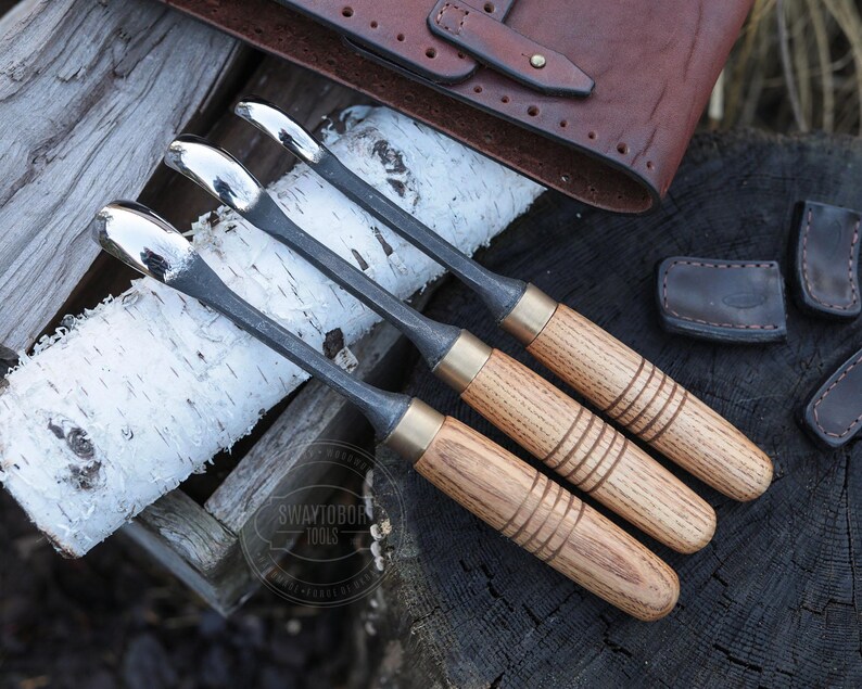 Może przedstawiać: Cztery dłuta do obr&oacute;bki drewna z polerowanymi metalowymi ostrzami i drewnianymi uchwytami. Uchwyty są jasnobrązowe z trzema ciemnobrązowymi paskami. Dłuta ułożone są na kawałku kory brzozy, z sk&oacute;rzanym etui na narzędzia w tle.