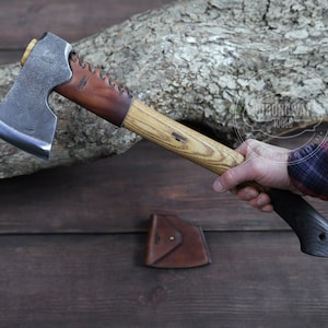 Könnte beinhalten: Axt mit Holzgriff und dunkler Metallklinge. Der Griff hat ein helles Holzmaserungsmuster und einen dunklen Griff. Eine Lederscheide ist sichtbar. Die Axt wird von einer Person gehalten.