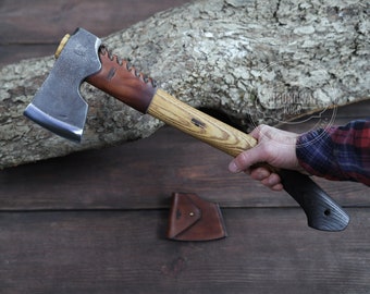 May include: Axe with a wooden handle and a dark metal blade. The handle has a light wood grain pattern and a dark grip. A leather sheath is visible. The axe is held by a person.