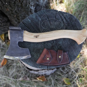 May include: A hand-forged axe with a wooden handle and a leather sheath. The axe has a curved blade and a rounded handle. The sheath is brown leather with a snap closure. The axe is sitting on a tree stump.