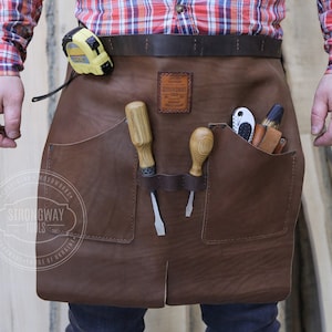 Könnte beinhalten: Braune Lederschürze mit mehreren Taschen, darunter eine große Tasche auf der rechten Seite. Die Schürze hat einen braunen Ledergürtel und ein gelbes Maßband, das am Gürtel befestigt ist. Die Schürze hat einen braunen Lederfleck mit dem Text "Strongway Tools" in Weiß.