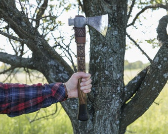 May include: A hand holds a  handmade axe with a wooden handle and a leather wrap. The axe head is made of metal and has a  distinctive shape. The handle is decorated with a geometric pattern. The axe is held against a tree trunk.