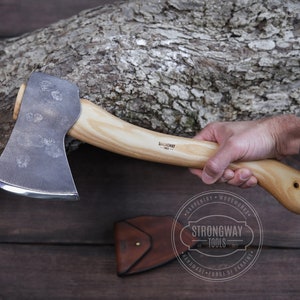 May include: A hand holds a  forged steel axe with a wooden handle. The axe has a  blackened finish and a  slightly curved blade. The handle is  light brown and has the text "Strongway Tools"  engraved on it. The axe is  resting on a  wooden surface.