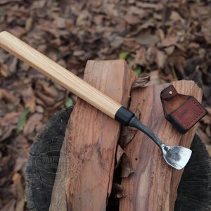 May include: A wooden handled carving tool with a black metal head and a curved blade. The tool is resting on a piece of wood with a leather sheath beside it.