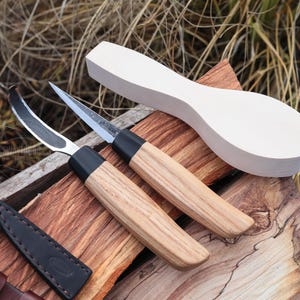 Puede incluir: Dos cuchillos para tallar madera con hojas curvas y mangos de madera, junto con una cuchara de madera en blanco y una funda de cuero. Los cuchillos tienen collares negros y están sobre una superficie de madera. La cuchara está sin terminar.