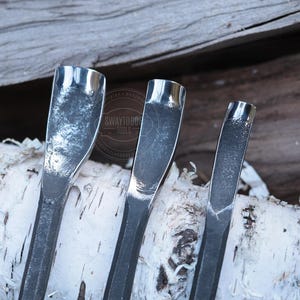 Może przedstawiać: Zbliżenie trzech metalowych narzędzi do rzeźbienia z zakrzywionymi ostrzami. Narzędzia są srebrne i mają teksturowany wygląd. Narzędzia są ułożone na kawałku kory brzozy, z drewnianym tłem. Narzędzia są oznaczone tekstem "SWAYTOBO TOOLS".