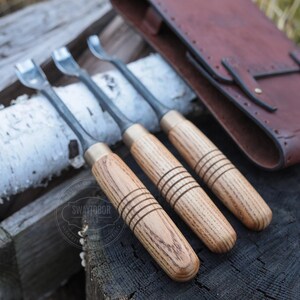 Może przedstawiać: Trzy dłuta do rzeźbienia w drewnie z metalowymi ostrzami i mosiężnymi tulejami. Rękojeści wykonane są z jasnego drewna z grawerowanymi liniami. W tle brązowe sk&oacute;rzane etui na narzędzia. Narzędzia leżą na drewnianej powierzchni.