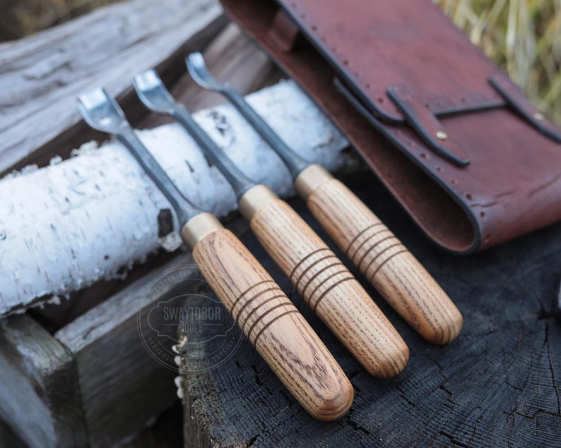 Może przedstawiać: Trzy dłuta do rzeźbienia w drewnie z metalowymi ostrzami i mosiężnymi tulejami. Rękojeści wykonane są z jasnego drewna z grawerowanymi liniami. W tle brązowe sk&oacute;rzane etui na narzędzia. Narzędzia leżą na drewnianej powierzchni.