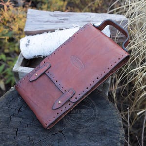 Może przedstawiać: Brązowa sk&oacute;rzana pochwa na n&oacute;ż z uchwytem, dwoma paskami z klamrami i ozdobnymi przeszyciami. Pochwa jest prostokątna i wydaje się być ręcznie robiona. Tło obejmuje drewno i liście.