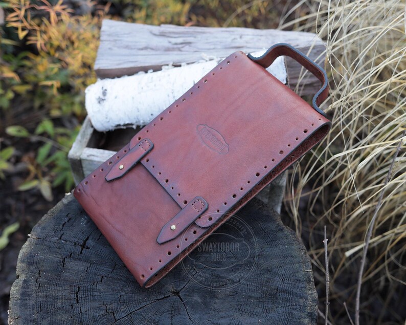 Może przedstawiać: Brązowa sk&oacute;rzana pochwa na n&oacute;ż z uchwytem, dwoma paskami z klamrami i ozdobnymi przeszyciami. Pochwa jest prostokątna i wydaje się być ręcznie robiona. Tło obejmuje drewno i liście.