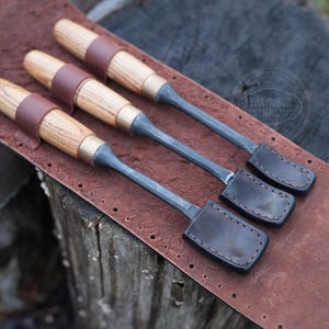 Może przedstawiać: Trzy dłuta do obr&oacute;bki drewna z drewnianymi uchwytami, mosiężnymi akcentami i sk&oacute;rzanymi paskami, spoczywające na brązowej sk&oacute;rzanej powierzchni. Dłuta mają ciemne metalowe ostrza i są ułożone w rzędzie. Sk&oacute;ra ma szytą obw&oacute;dkę.