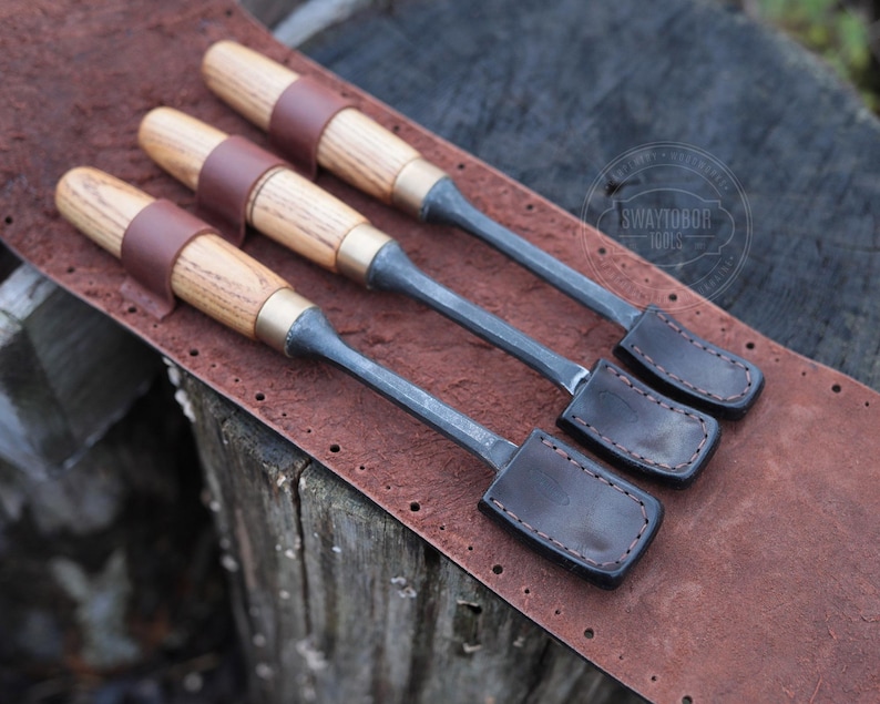 Może przedstawiać: Trzy dłuta do obr&oacute;bki drewna z drewnianymi uchwytami, mosiężnymi akcentami i sk&oacute;rzanymi paskami, spoczywające na brązowej sk&oacute;rzanej powierzchni. Dłuta mają ciemne metalowe ostrza i są ułożone w rzędzie. Sk&oacute;ra ma szytą obw&oacute;dkę.