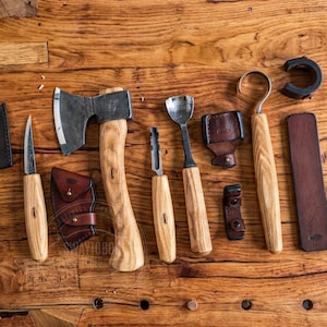 May include: A collection of woodworking tools, including an axe, knives, and a spoon carving tool, arranged on a wooden surface. The tools have wooden handles and leather sheaths. The axe head is silver, and the handles are light-colored wood.