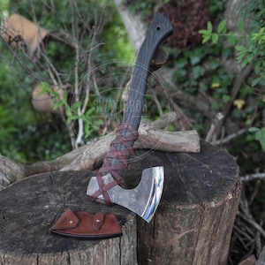 May include: A hand axe with a dark, textured wooden handle wrapped in brown leather lacing. The metal blade has a polished, curved edge. A separate brown leather sheath with two brass rivets lies on the surface next to the axe head.