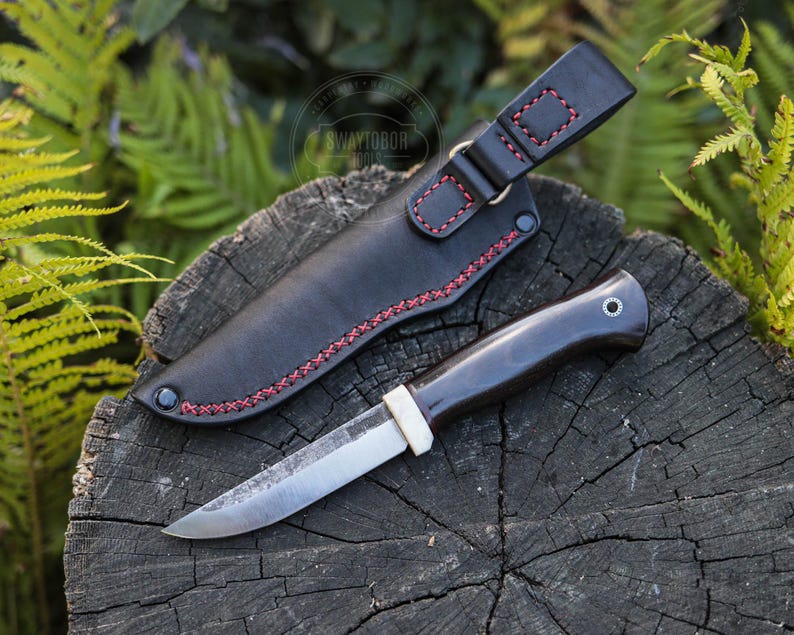 May include: A handcrafted knife with a dark handle and a silver blade, resting on a wooden surface. The knife is accompanied by a black leather sheath with red stitching. The overall design suggests a focus on craftsmanship and outdoor utility.