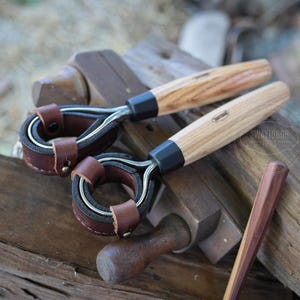 May include: Two wood-handled leather tools with metal rings and brown leather accents. The handles are light-colored wood with a dark band near the metal. The leather is a rich brown color. A small wooden tool is also present.