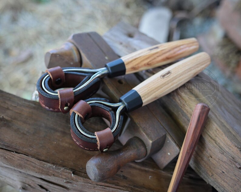May include: Two wood-handled leather tools with metal rings and brown leather accents. The handles are light-colored wood with a dark band near the metal. The leather is a rich brown color. A small wooden tool is also present.