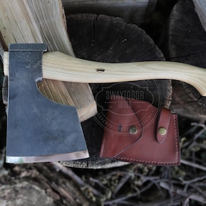 May include: A hand axe with a dark, forged steel head and a light-colored wooden handle. The axe features a brown leather sheath with a metal snap closure. The text "SWAYTOBOR TOOLS" is visible on the leather sheath and the wooden handle.