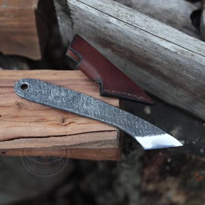 Op de afbeelding: Een klein, handgesmeed mes met een getextureerd, grijs metalen blad en een bruine leren schede. Het mes ligt op een stuk hout.