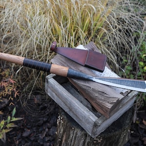 Può includere: Uno scalpello forgiato a mano con manico in legno e fodero in pelle. Lo scalpello ha una lama in metallo scuro e un manico in legno avvolto in pelle nera. Il fodero è marrone. Lo scalpello poggia su una scatola di legno.