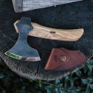 May include: A hand axe with a dark metal head and a light brown wood handle featuring the text "BEAVER" on the blade and handle. A brown leather sheath with a brass snap button is positioned next to the axe head, showing a small logo.