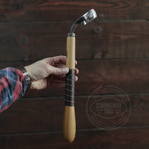 以下が含まれることがあります： 木製のハンドルに革が巻かれた金属製の刃が付いた彫刻用具。木製の背景に手によって保持されている。背景の円の中に「Strongway Tools」という文字が見える。